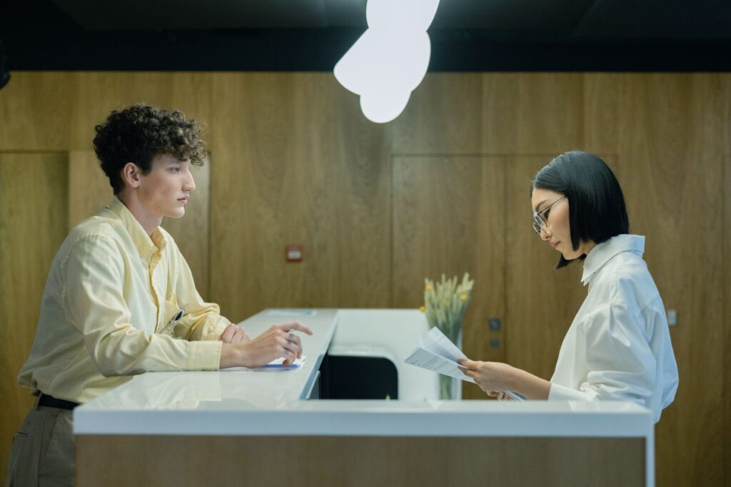 A young man and woman interacting at a modern office reception desk indoors.